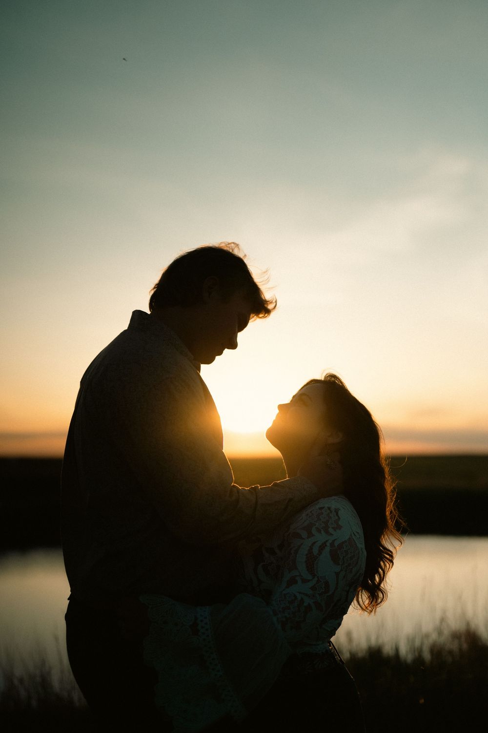 Silhouetted couple embraces against sunset sky with waterfront view and golden sunlight glowing behind them.