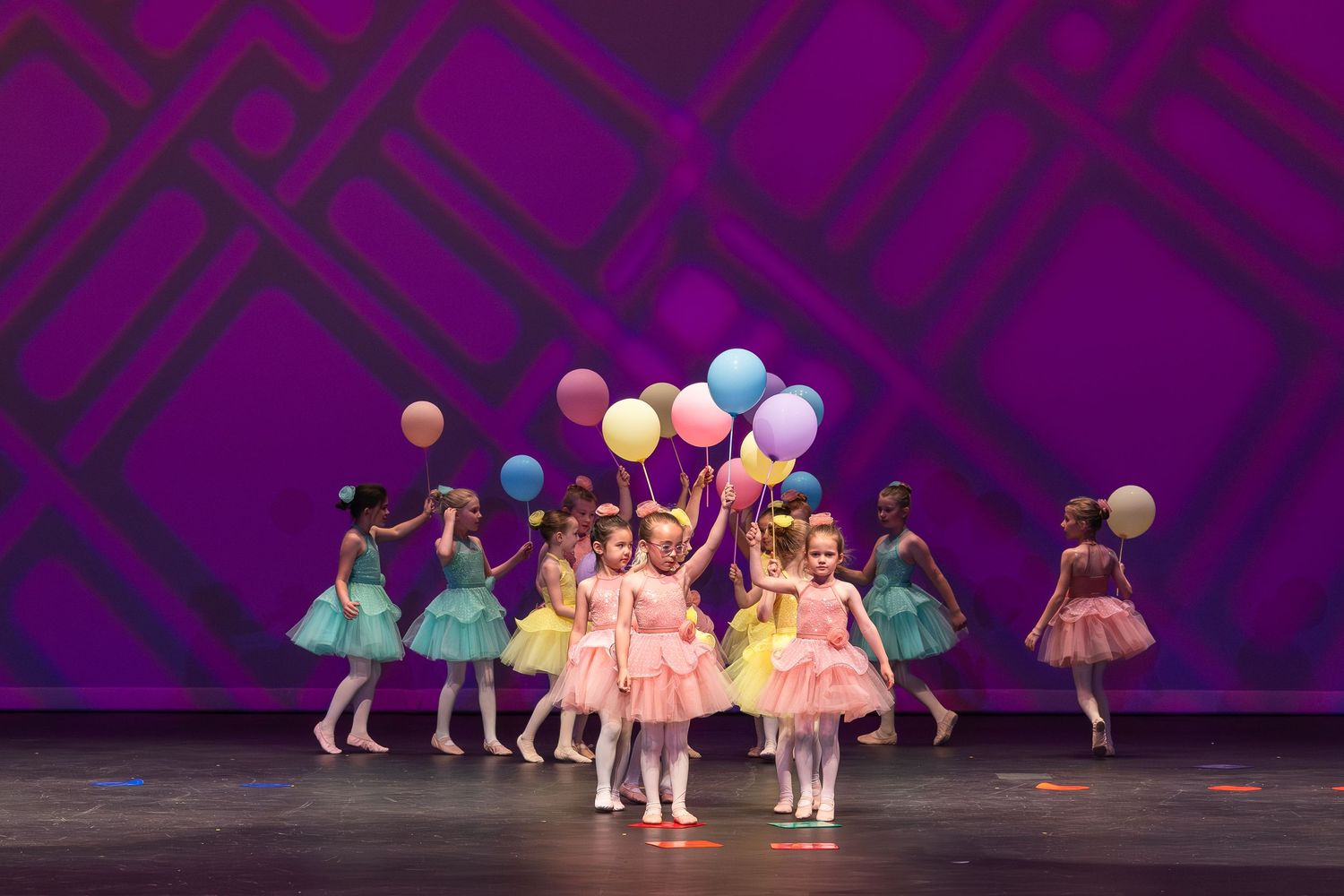Young ballet dancers in tutus and leotards perform on stage during a dance recital with purple lighting.
