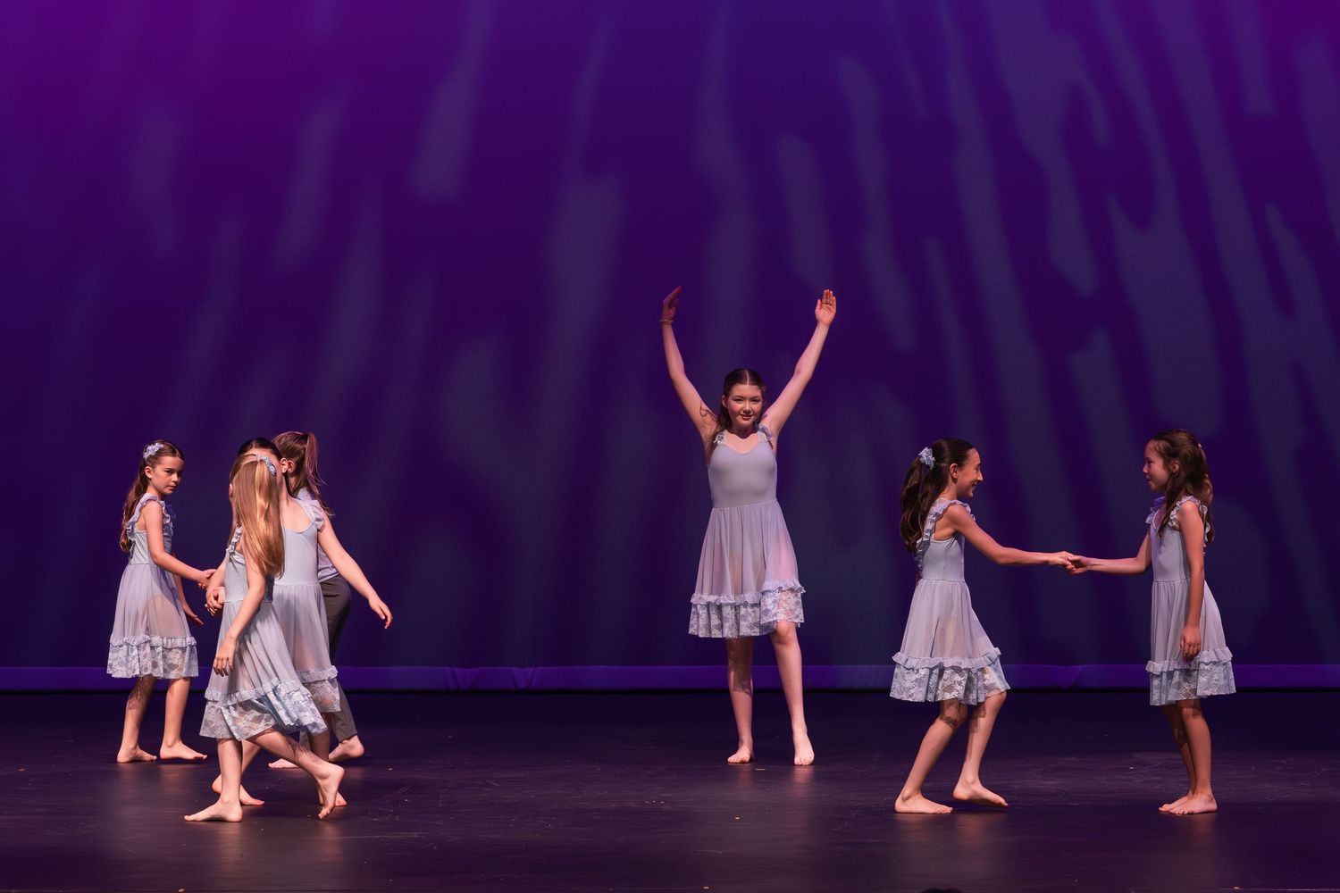Ballet dancers in white dresses perform a graceful sequence on a purple-lit stage during a dance recital.