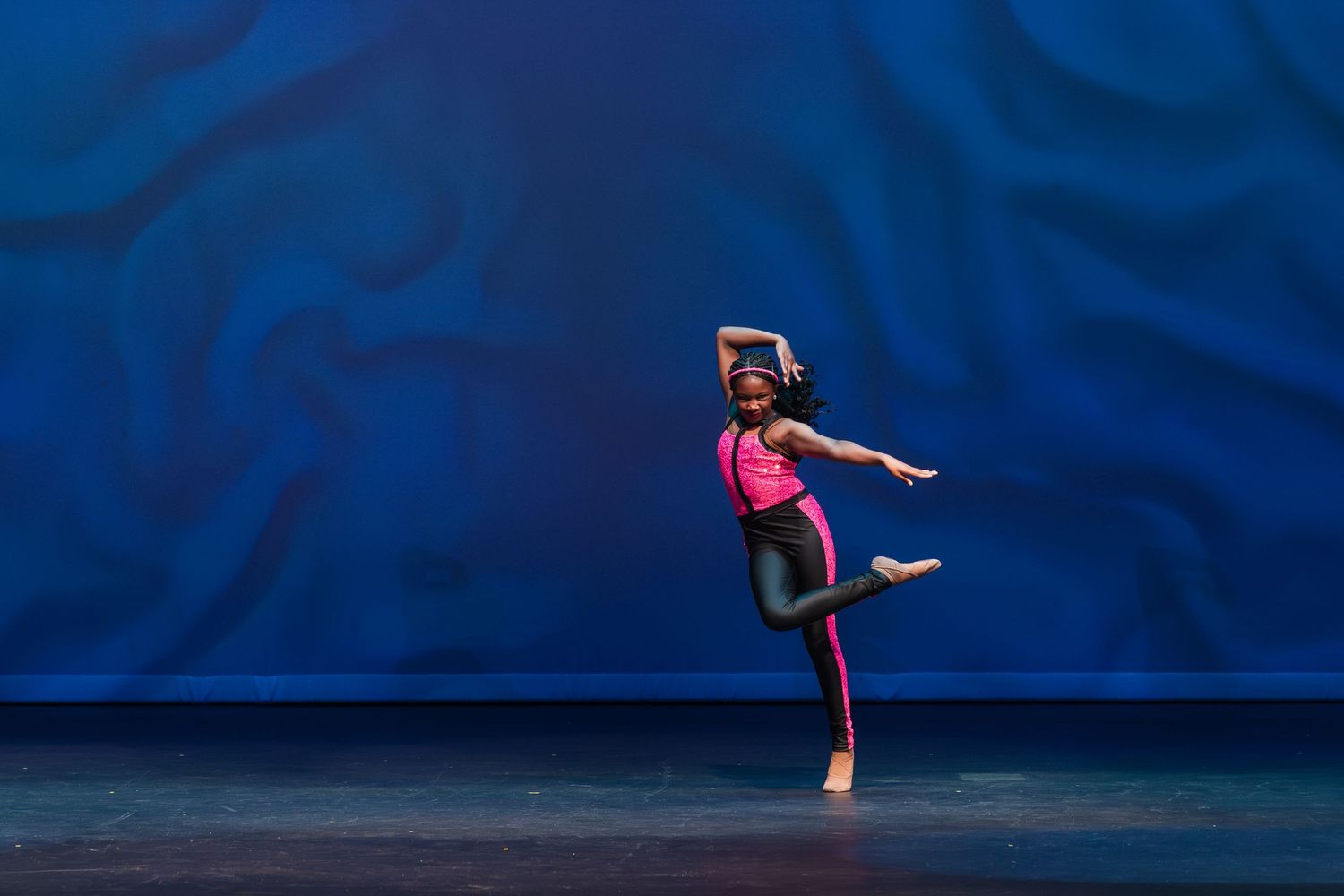 A graceful dancer in a black leotard performs an elegant leap against a deep blue stage backdrop.