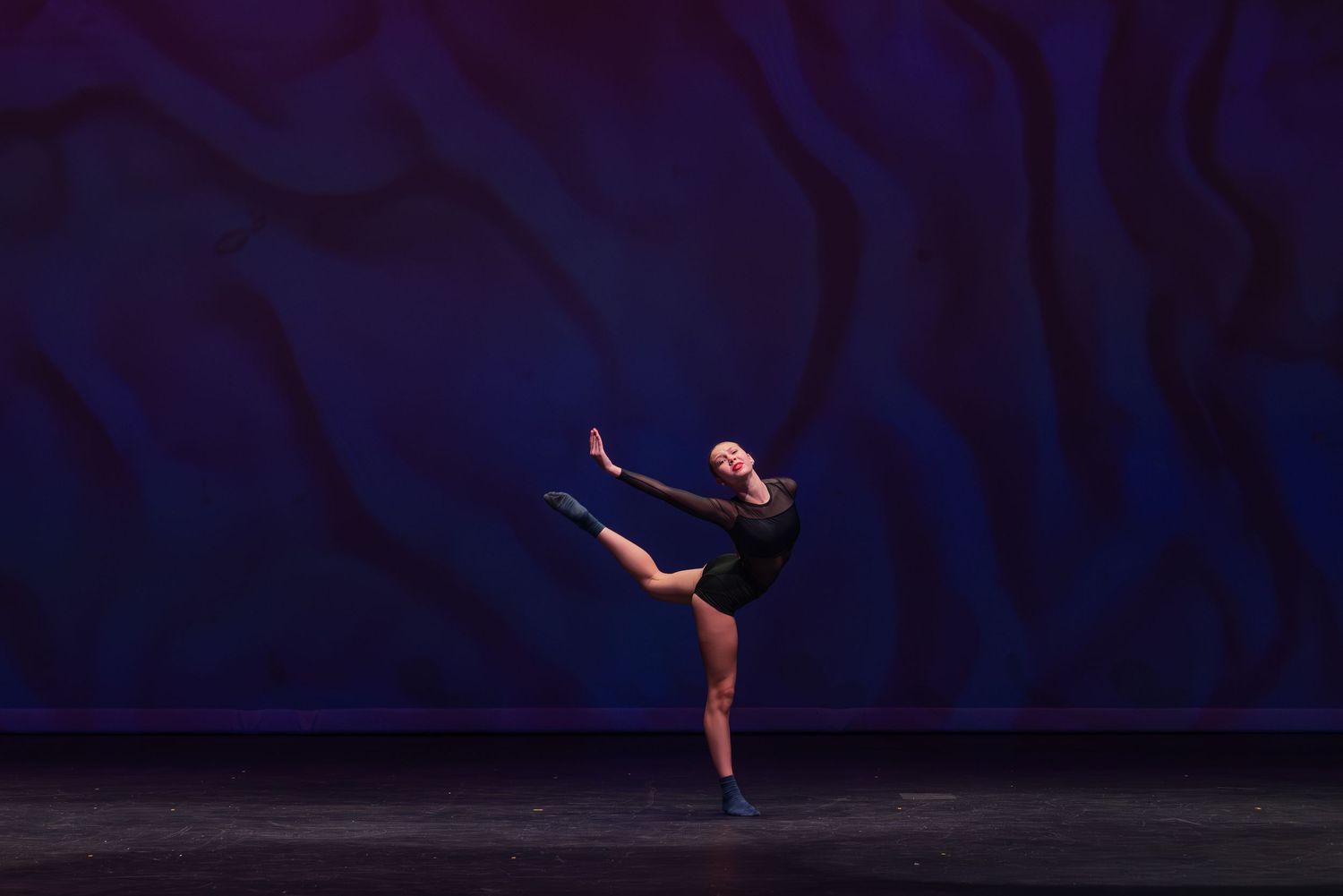 Ballet dancer in black leotard performs graceful arabesque pose against dark blue backdrop.