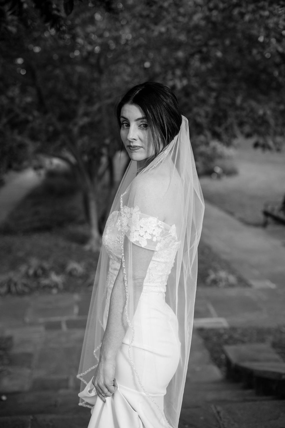 Black and white portrait of a bride in a flowing veil and off-shoulder wedding dress.