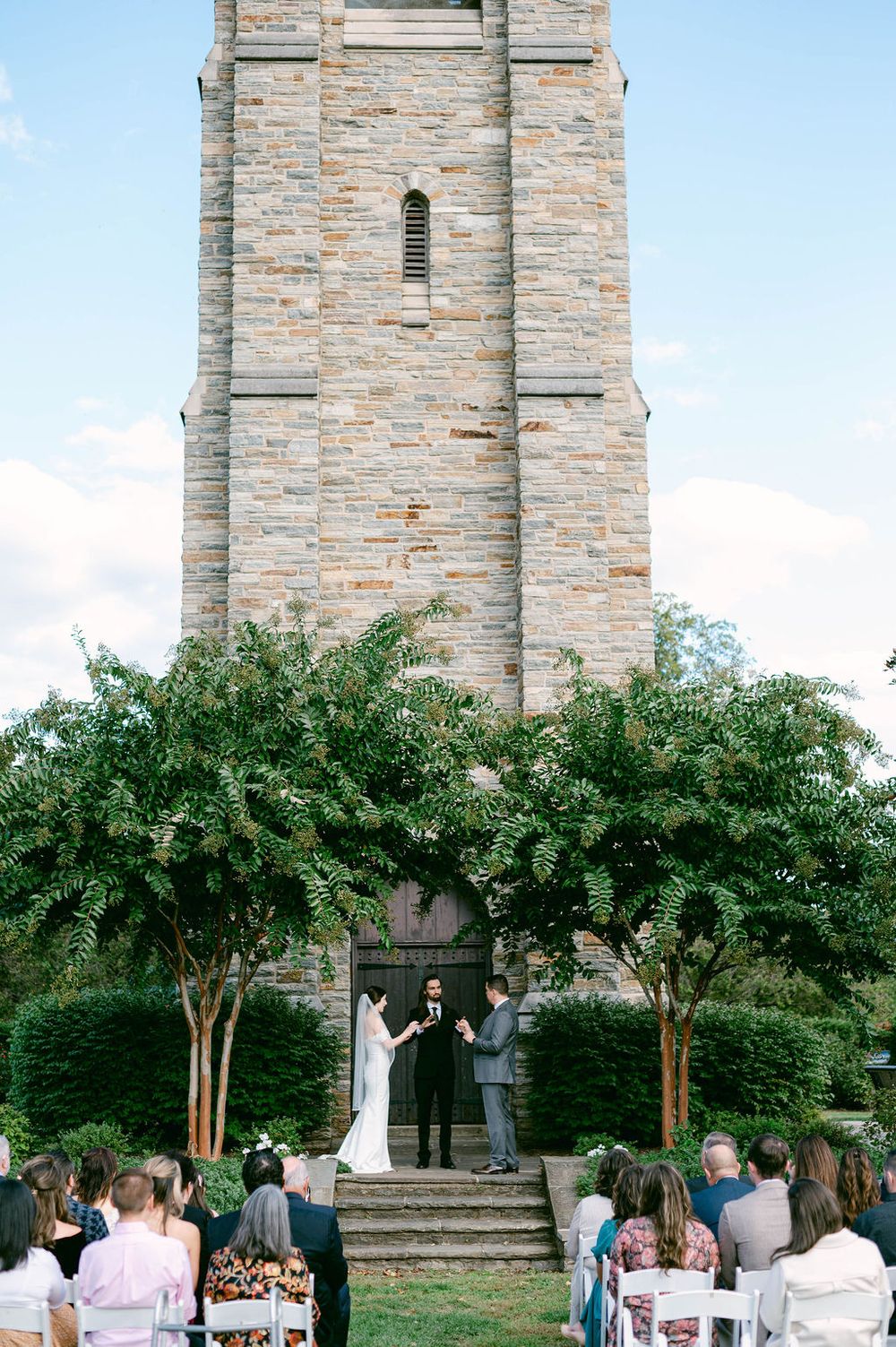 Outdoor wedding ceremony in front of a stone church tower with guests seated on white chairs surrounded by trees.