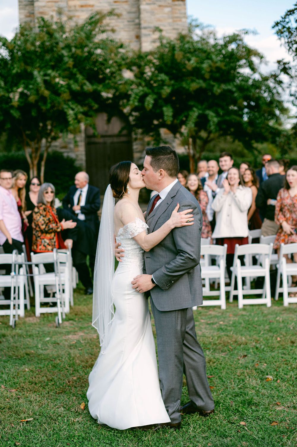 A couple shares their first kiss as newlyweds during an outdoor garden wedding ceremony with guests seated on white chairs.