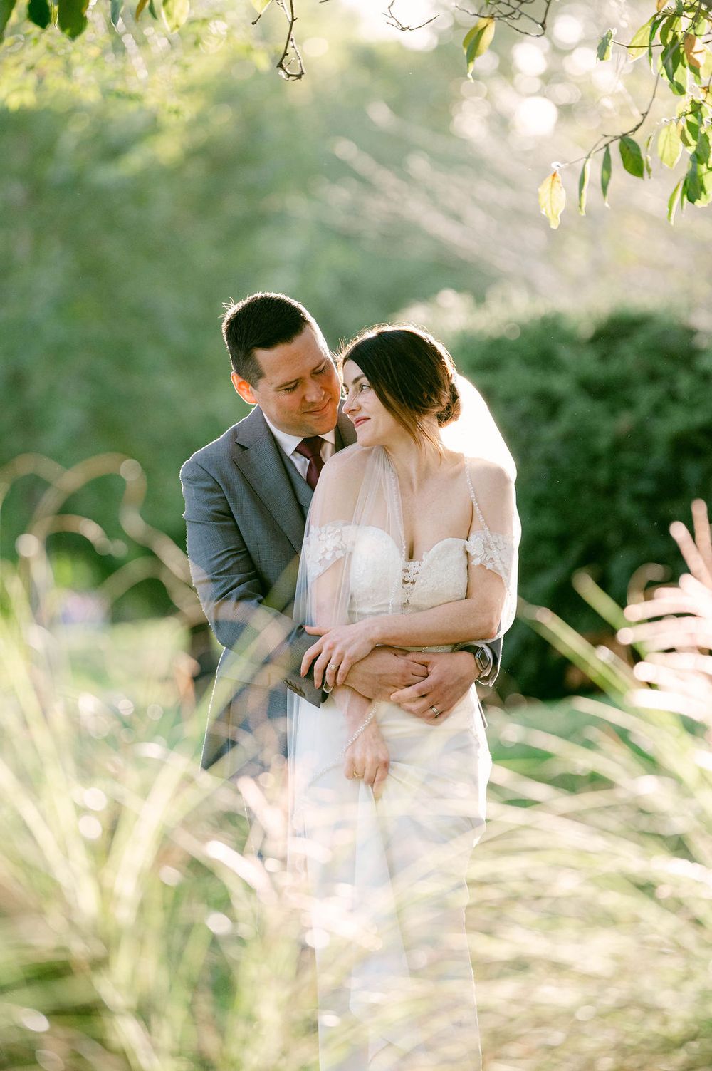 A romantic wedding moment captured in soft sunlight with greenery and bokeh in the background.