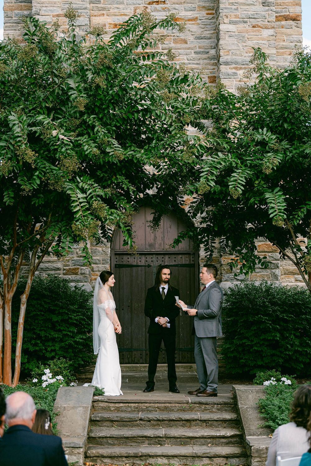 A wedding ceremony takes place on stone steps outside a wooden church door framed by lush green foliage.