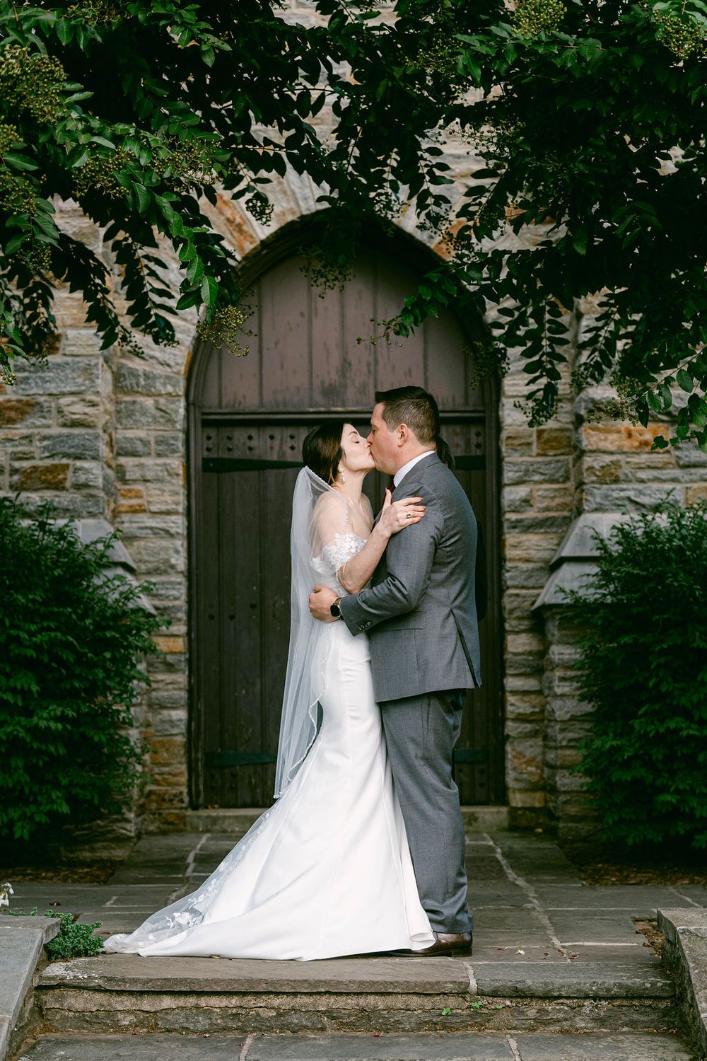 A romantic wedding couple embraces in front of an arched stone church doorway framed by green foliage.
