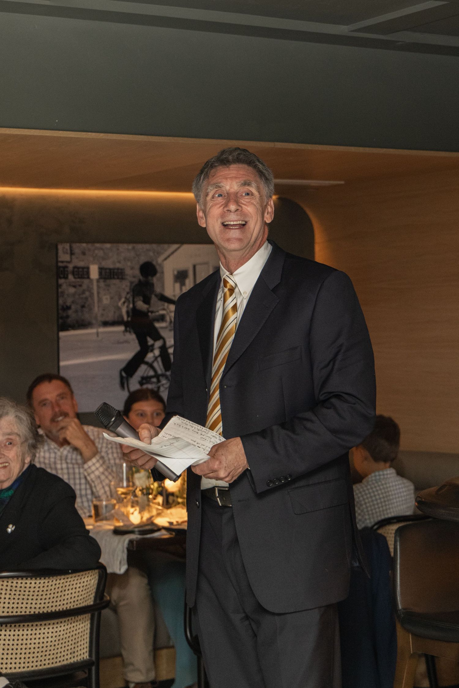 A person in a suit and striped tie speaks at a wedding reception in a restaurant setting.