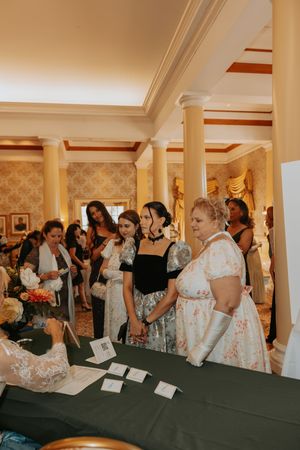Attendees gather around a table at an elegant indoor event with decorative columns and patterned wallpaper.