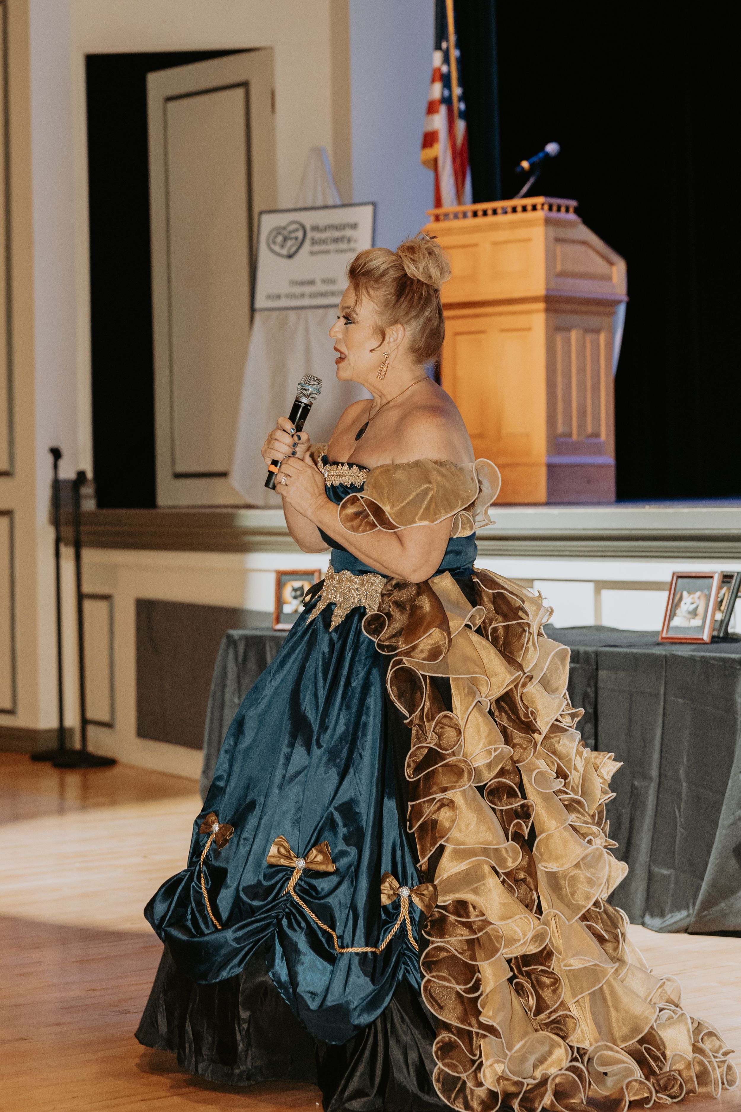 Performer in an elaborate blue and gold Victorian-style ball gown speaks at a podium on stage.
