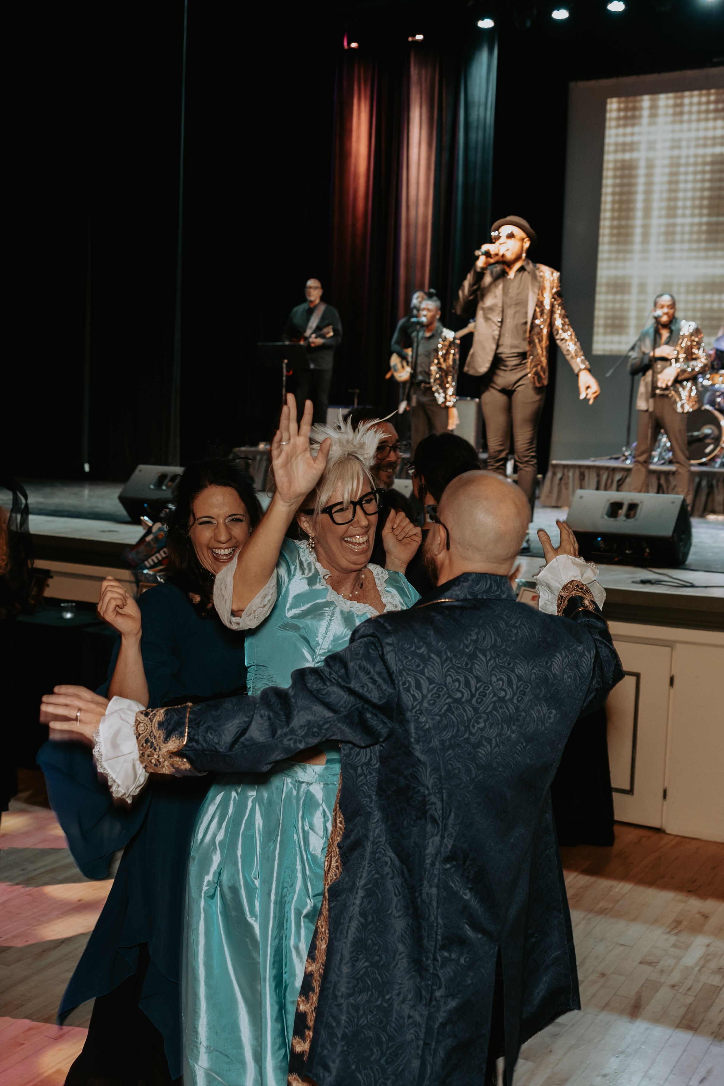 People dancing and celebrating at an indoor event while band performs on stage in background.