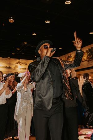 Performer in black attire and hat sings passionately while audience raises hands in celebration.