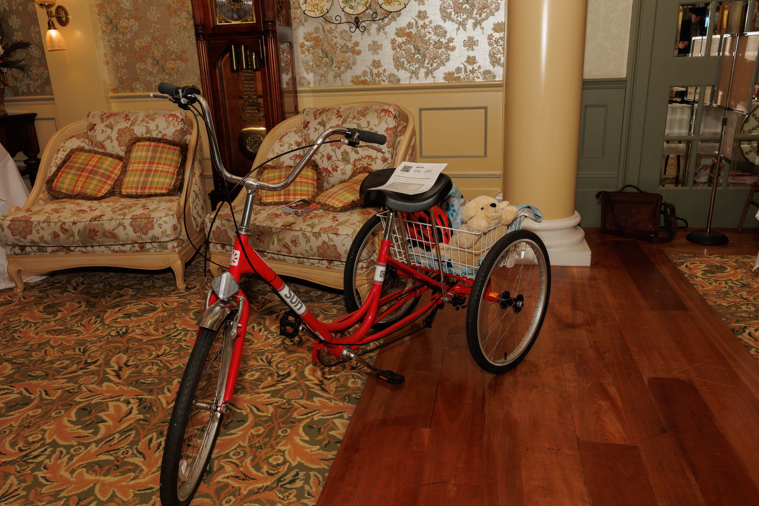 Red tricycle displayed in a room with ornate floral carpeting and beige walls.