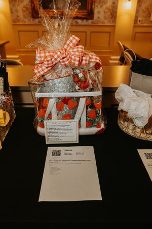 A gift basket wrapped in red gingham ribbon displayed on a black table with paperwork at a silent auction.