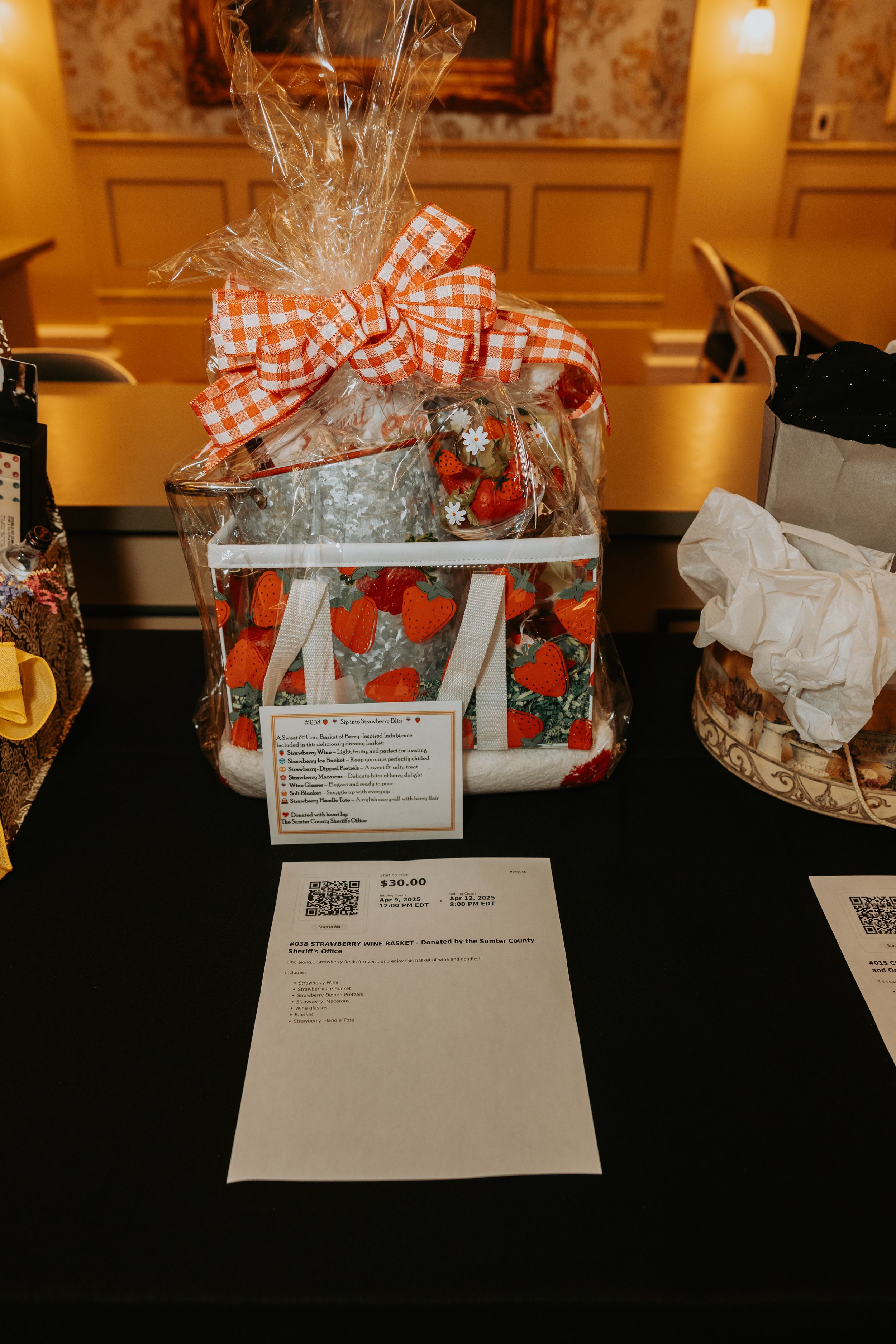 A gift basket wrapped in red gingham ribbon displayed on a black table with paperwork at a silent auction.