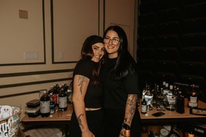 Two people stand together at a bar area with bottles and glassware displayed behind them.