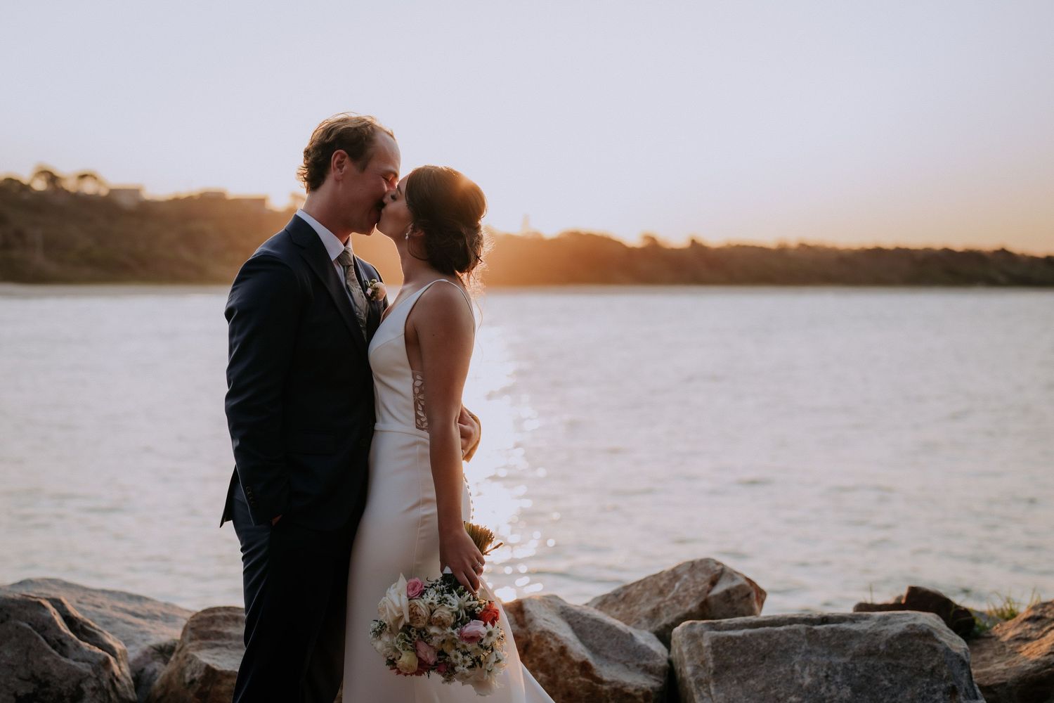 A romantic sunset wedding portrait of a couple standing on rocky coastline.