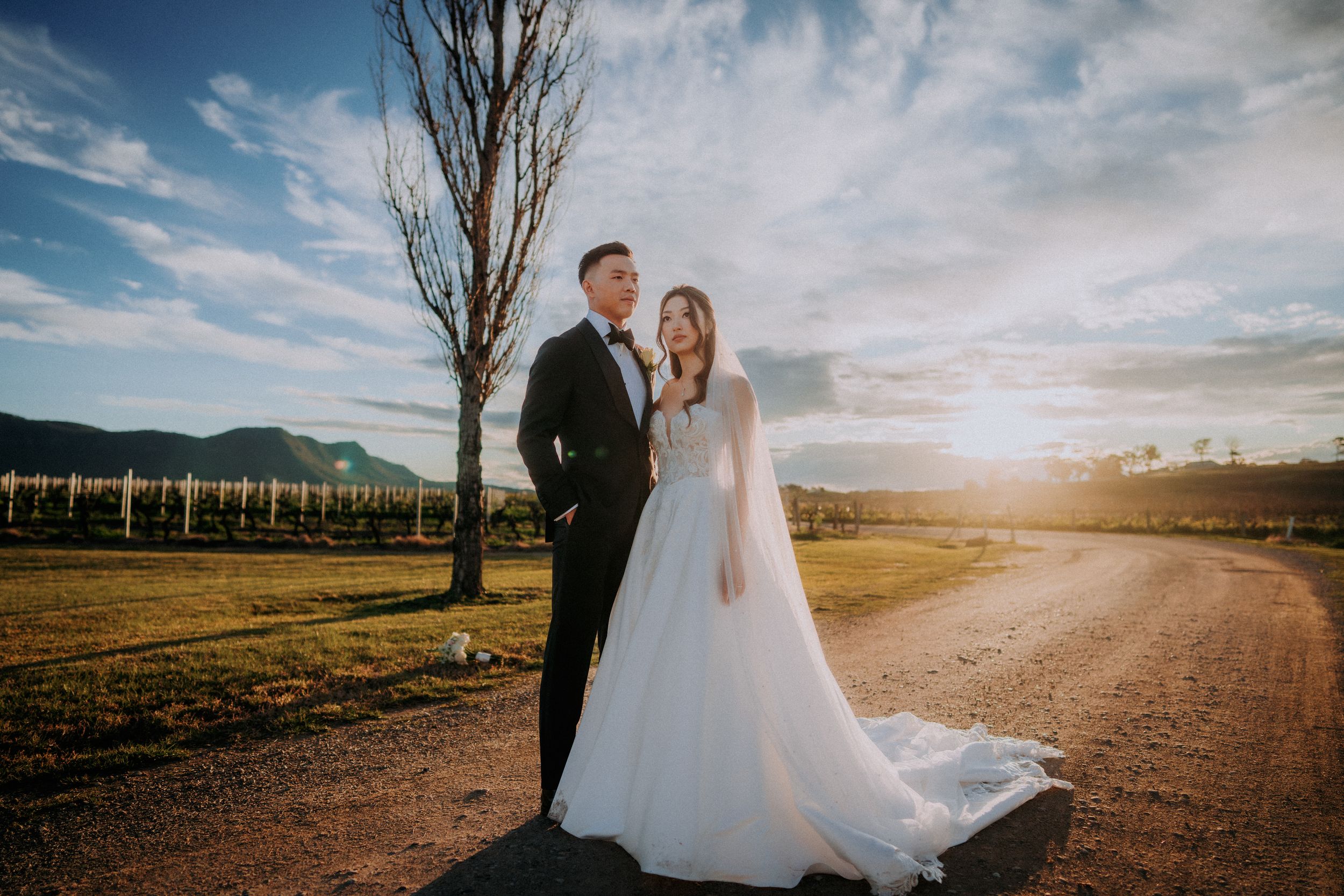 Newlyweds pose together in a scenic vineyard with dramatic clouds overhead.