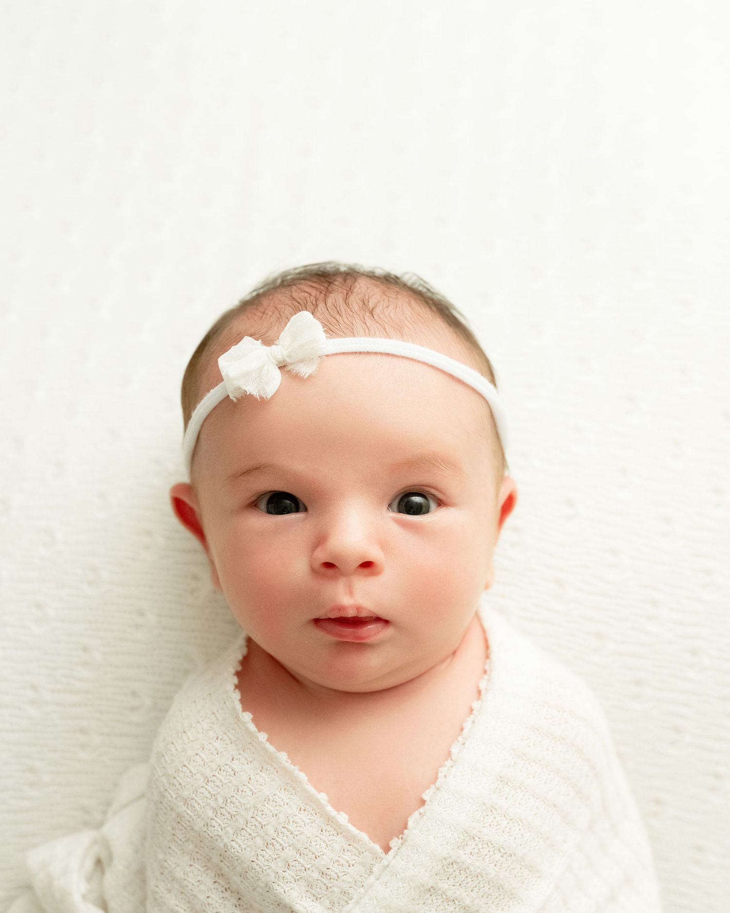 Baby wearing white lace dress and small floral headband smiles while lying on white background.