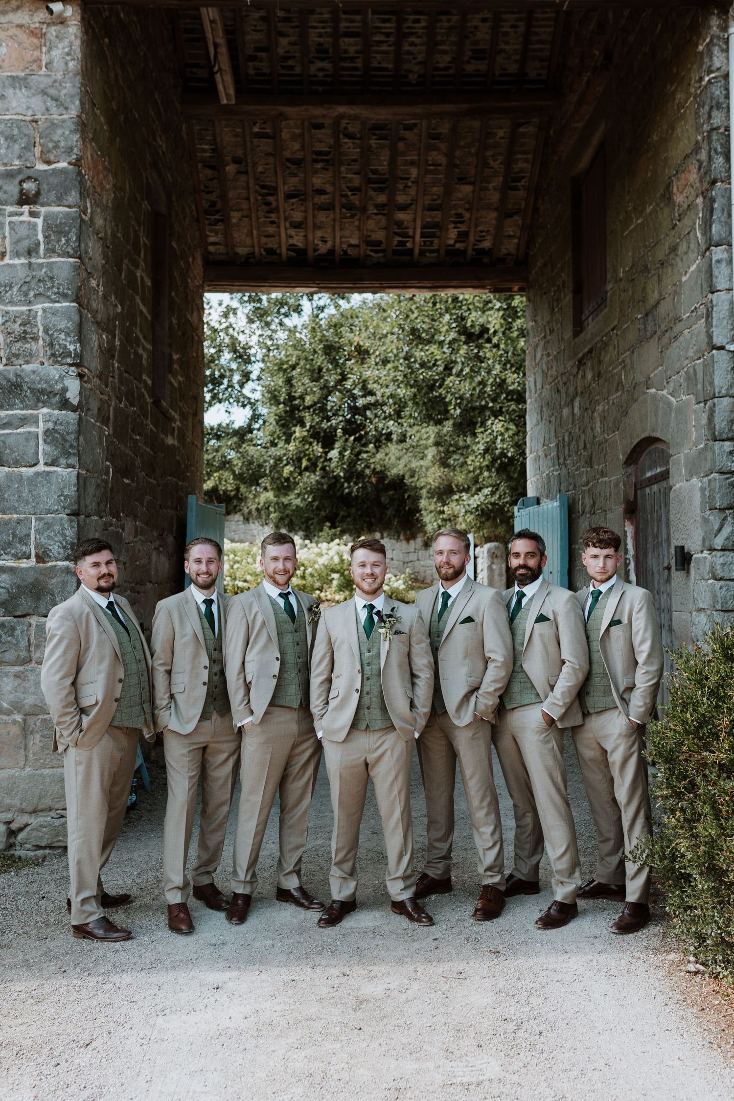 Groomsmen in matching tan suits and brown shoes lean against stone wall under covered walkway at wedding venue.