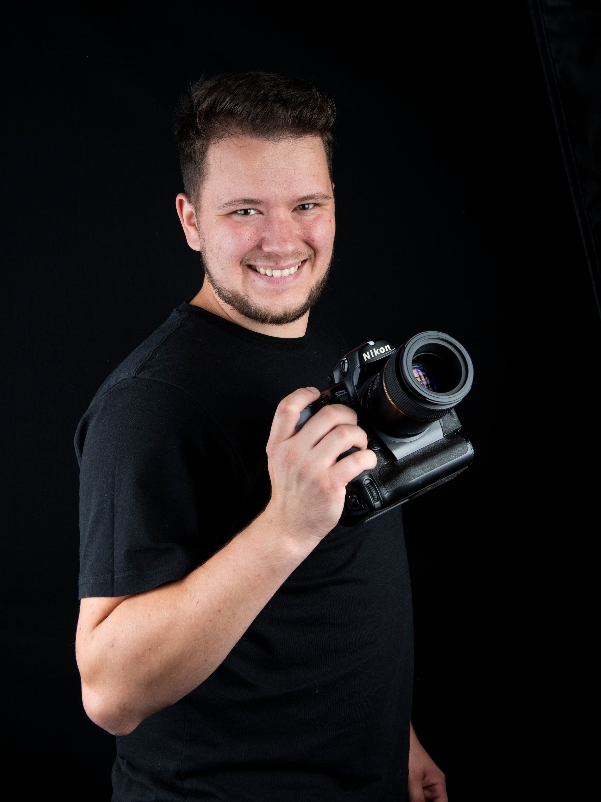 A photographer in a black t-shirt holds a professional DSLR camera against a dark background.