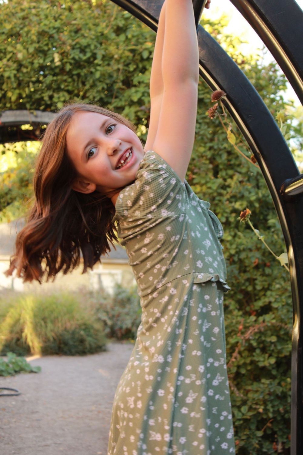 Young person in floral dress twirling and playing under an outdoor garden arch.