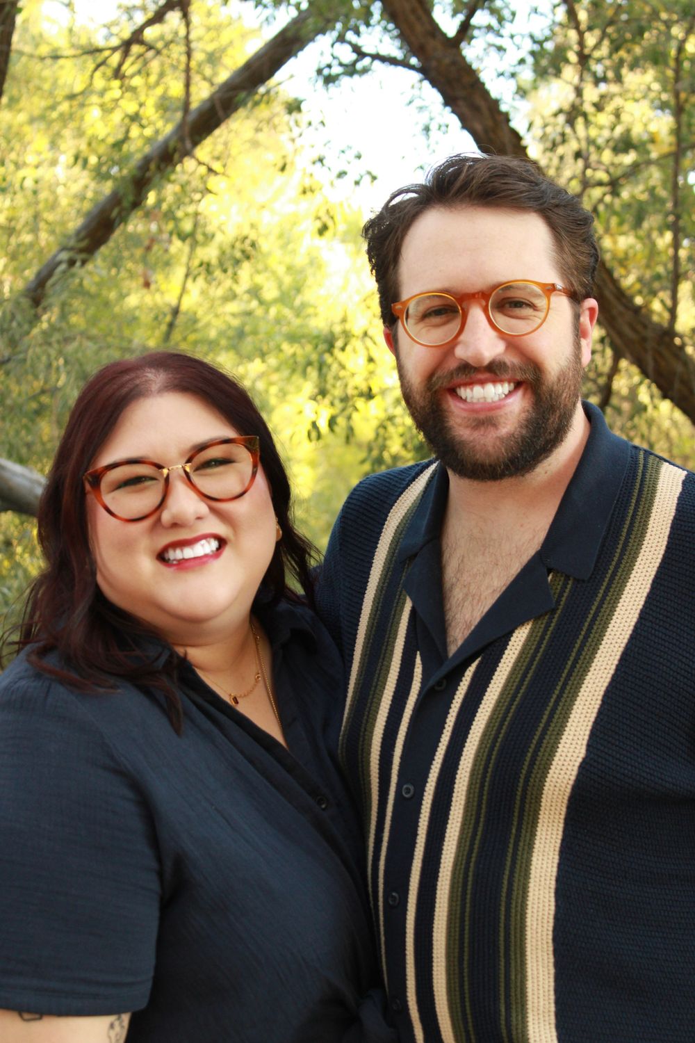 A couple in dark clothing shares joyful laughter while posing together outdoors with sunlit trees in the background.