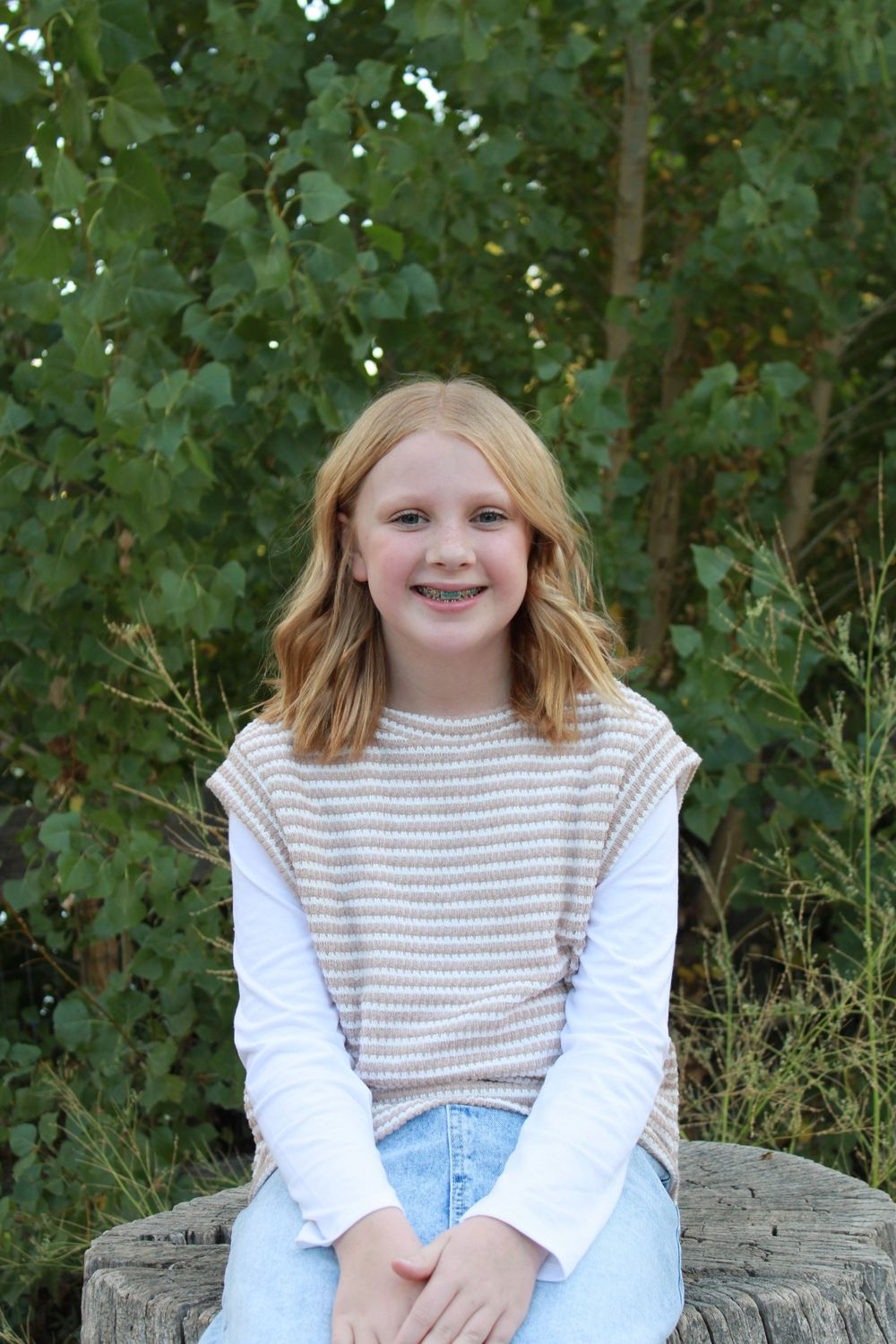 A young girl in a striped top sits on a stone wall outdoors smiling at the camera.