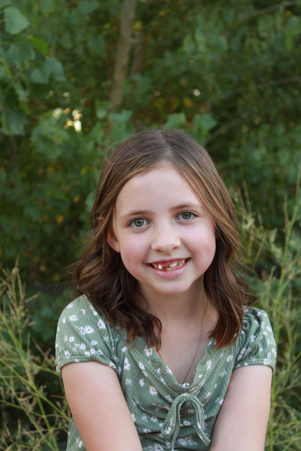 A young child in a green floral dress smiles at the camera against a leafy outdoor background.