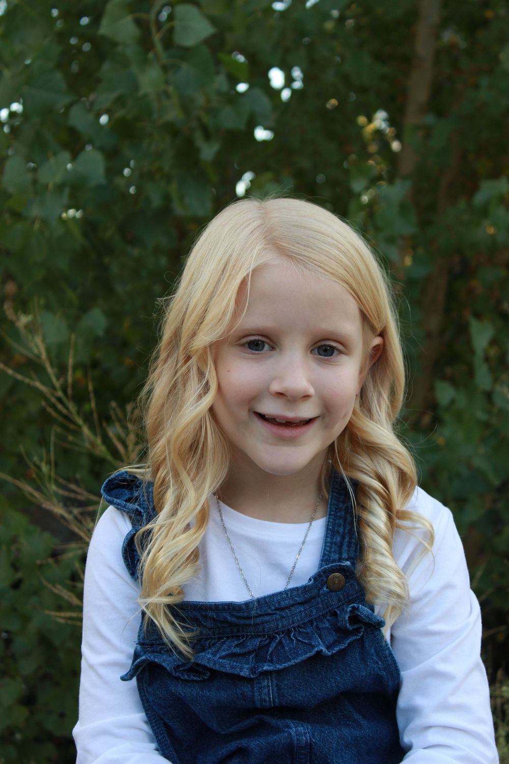 Young child in denim overalls and white shirt smiling during outdoor portrait session.