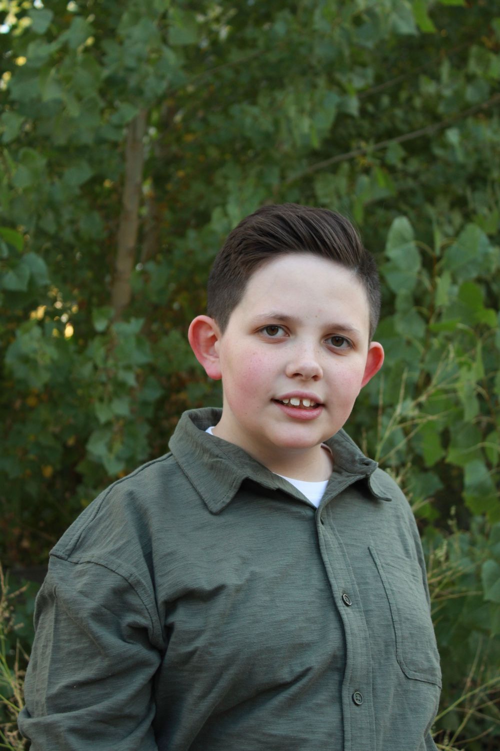 A youth wearing an olive green collared shirt poses for a portrait outdoors.