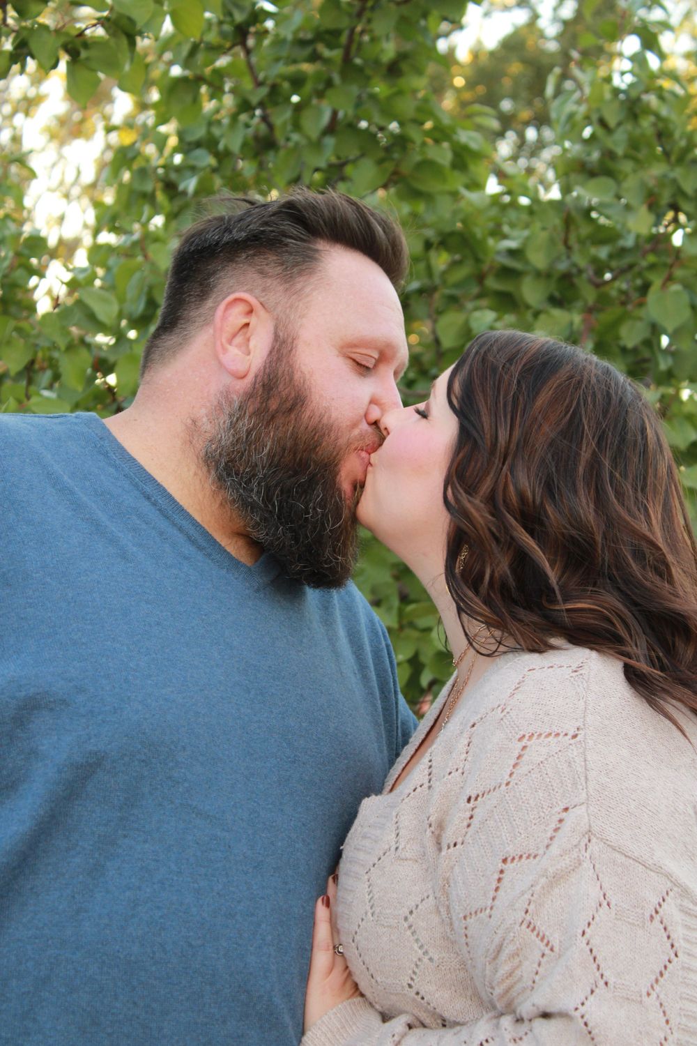 A romantic couple shares a tender kiss outdoors with green foliage in the background.