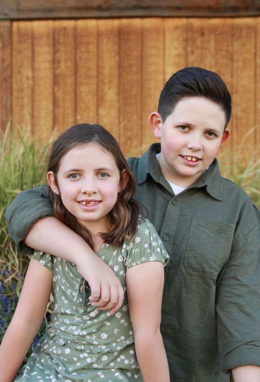 Two children in casual attire pose together outdoors against a wooden fence background.