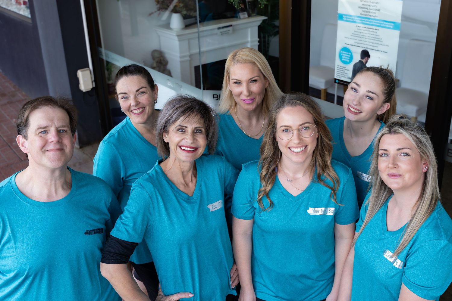Seven women in matching teal uniforms smile together inside a modern healthcare clinic.