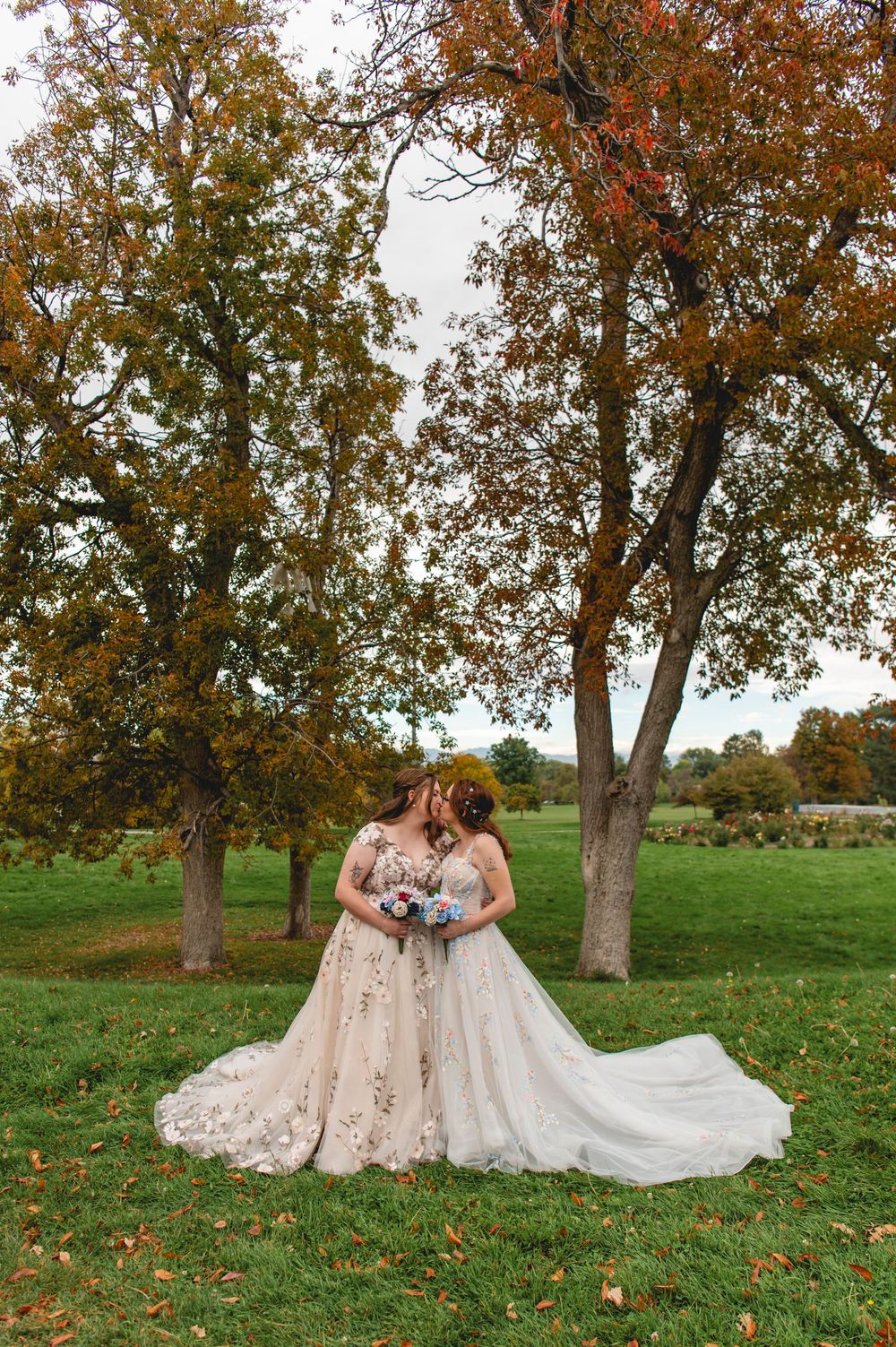 Two brides in flowing white lace gowns share an intimate moment in a fall orchard setting with autumn leaves.