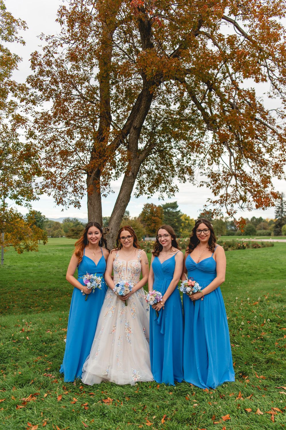 Wedding party wearing blue bridesmaid dresses and white wedding gown stand together on grass with fall foliage overhead.