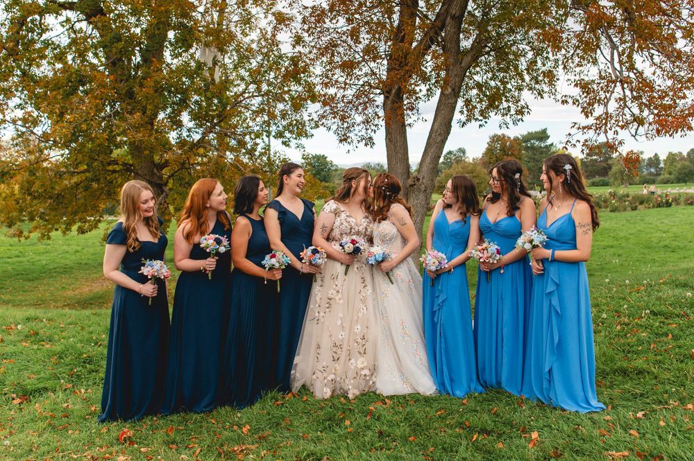 Wedding party group sits together on grass with bridesmaids in coordinating blue dresses during fall season.