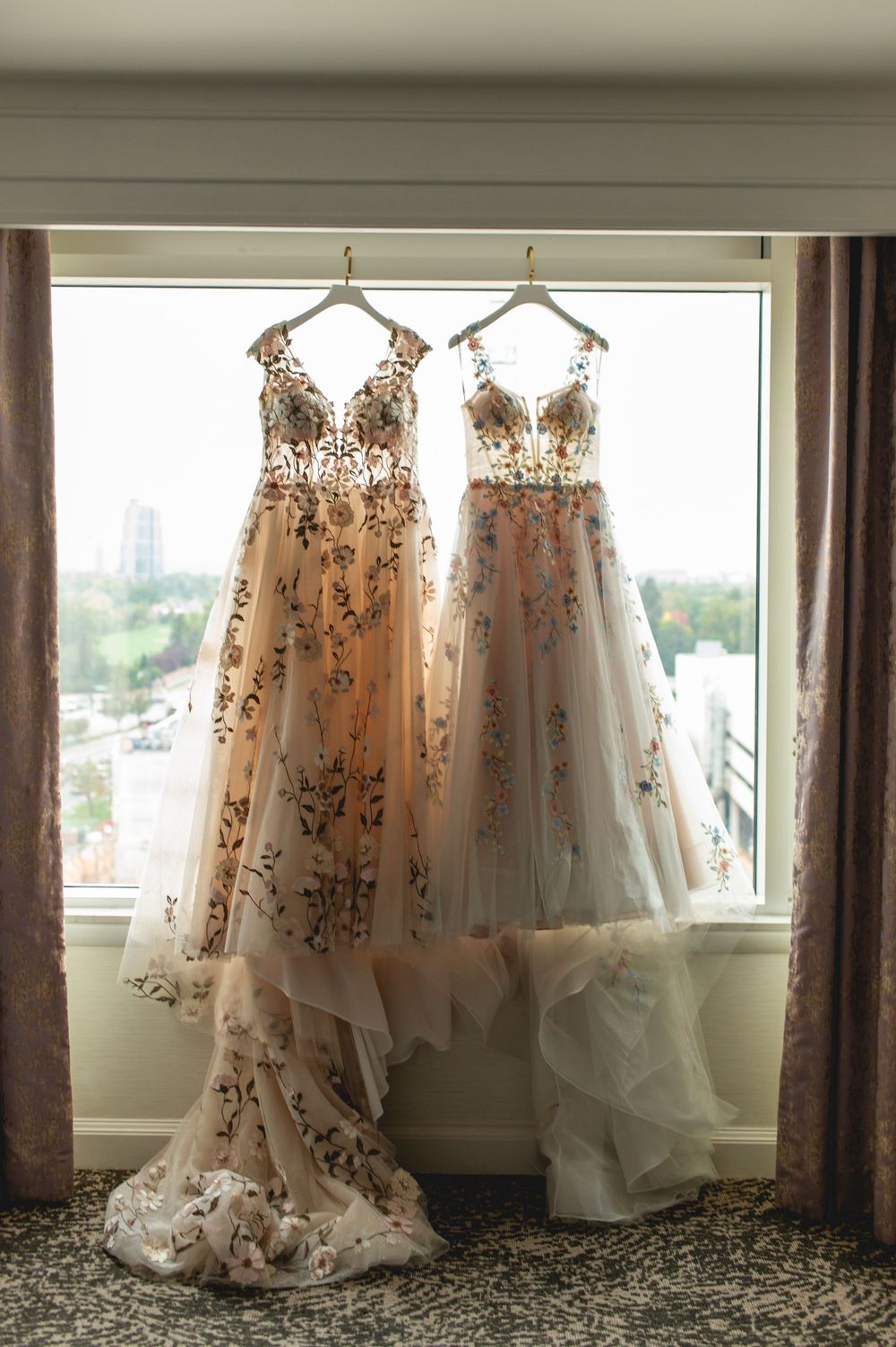 Two elegant floral embroidered wedding gowns hanging by window with sheer tulle and delicate beaded details.