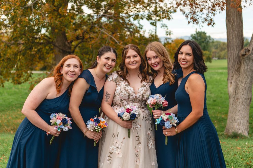 A group in navy blue and beige dresses poses together outdoors under autumn trees with colorful bouquets.