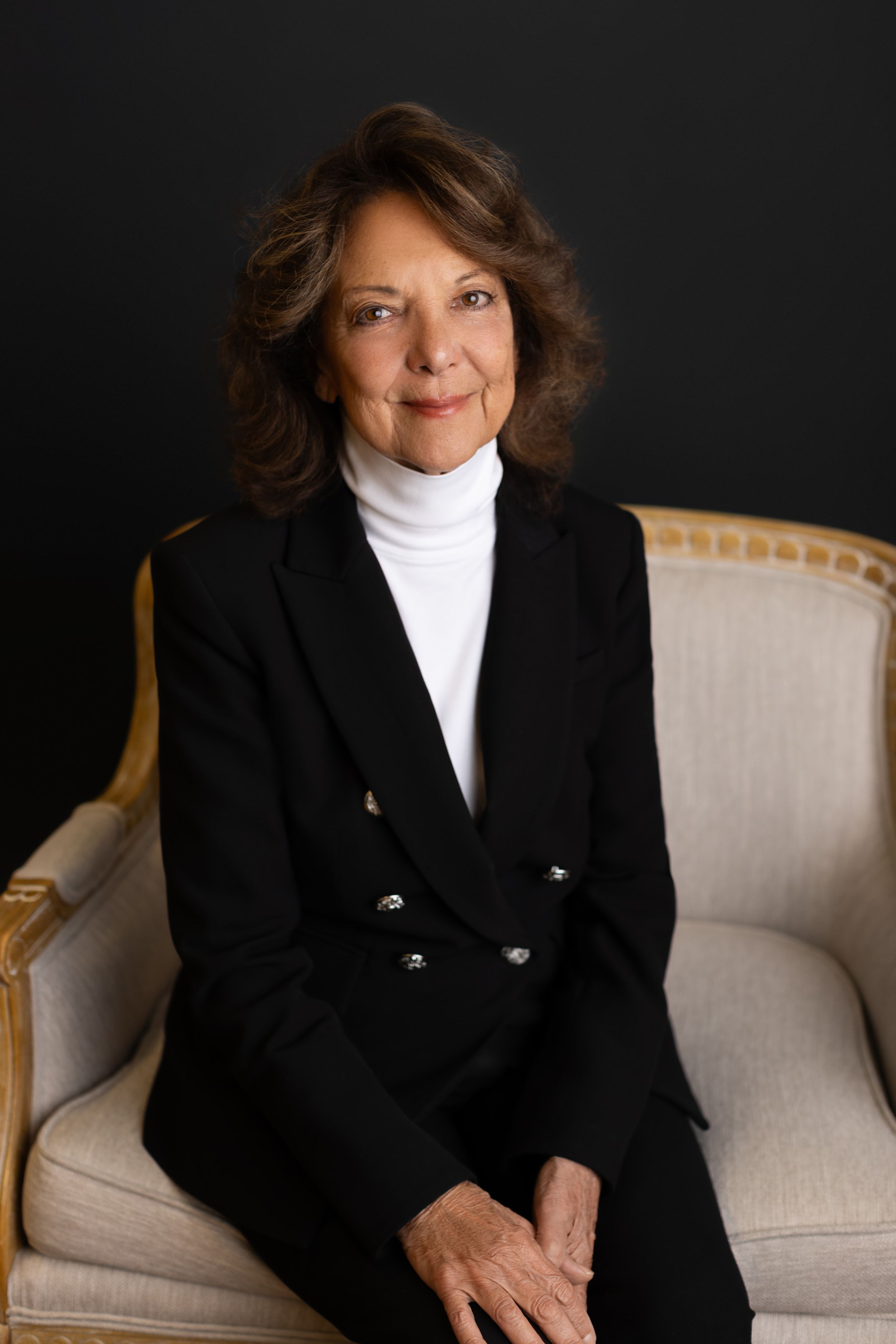 Professional portrait of someone wearing a black blazer and white turtleneck while seated in an elegant cream-colored chair. NJ headshots
