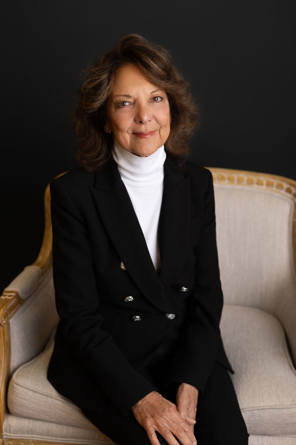 Professional portrait of someone wearing a black blazer and white turtleneck while seated in an elegant cream-colored chair. NJ headshots