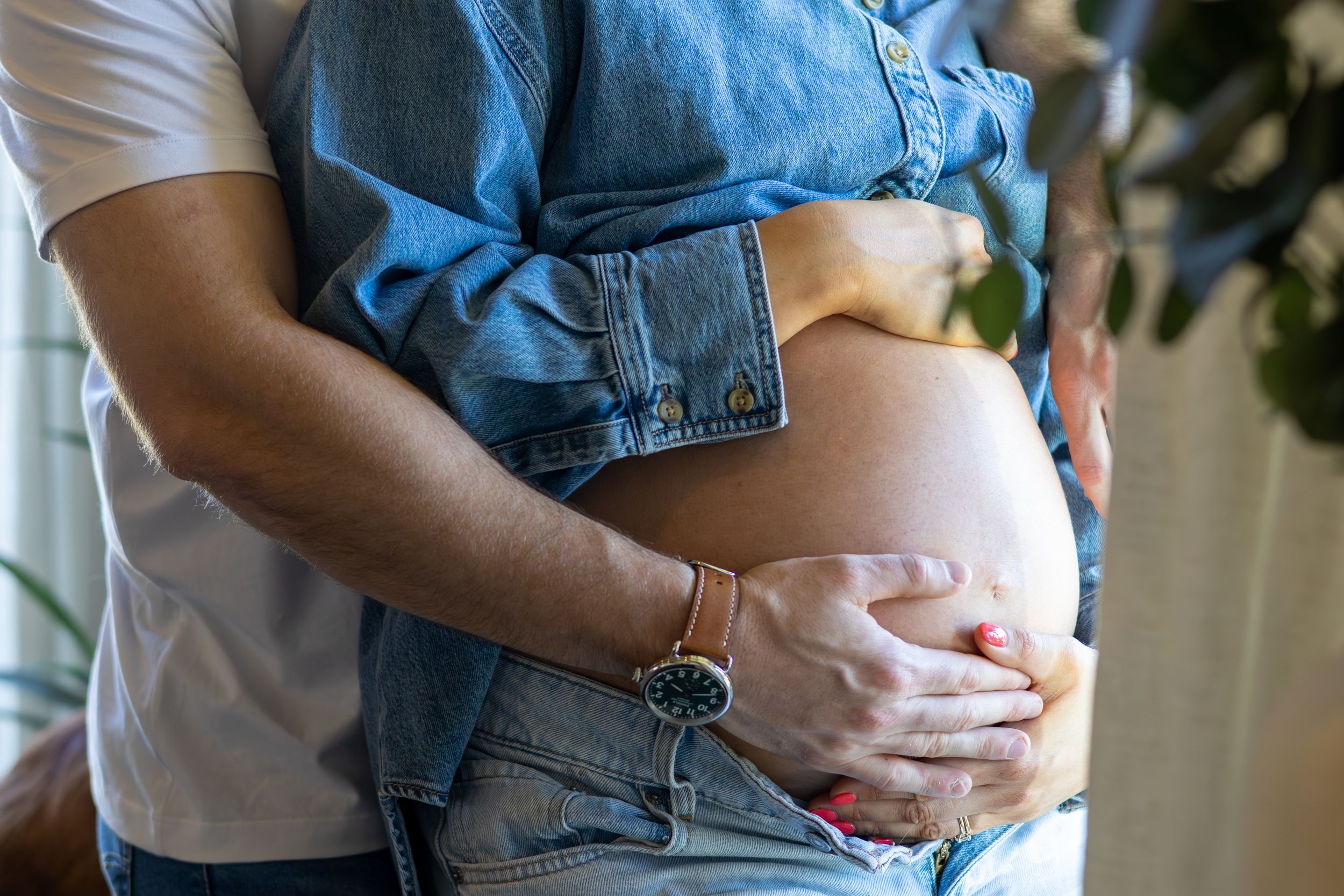 Partner gently embracing pregnant woman from behind, lovingly cradling her bare baby bump indoors.
