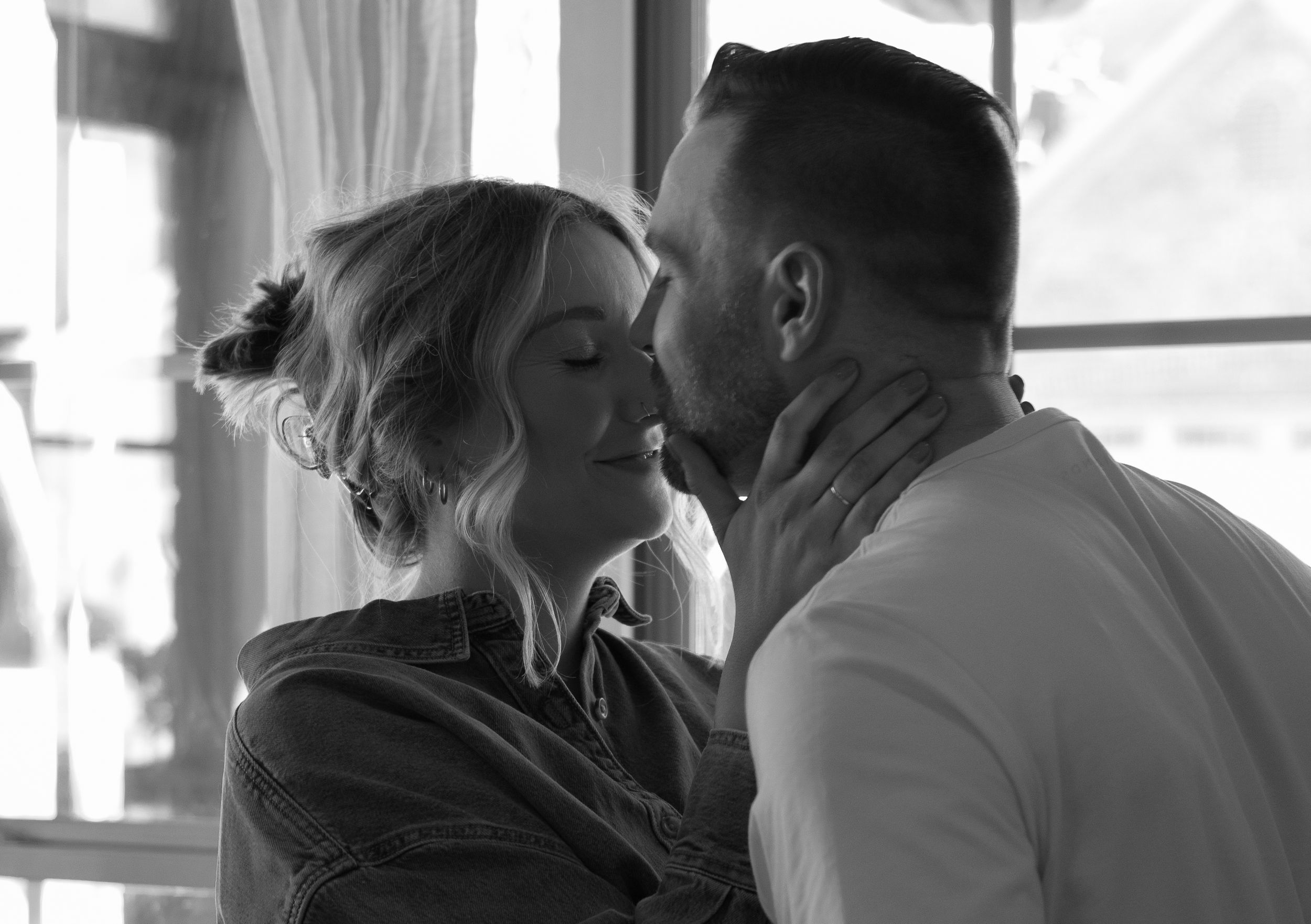 Romantic black and white photo of couple sharing a kiss near a sunlit window.