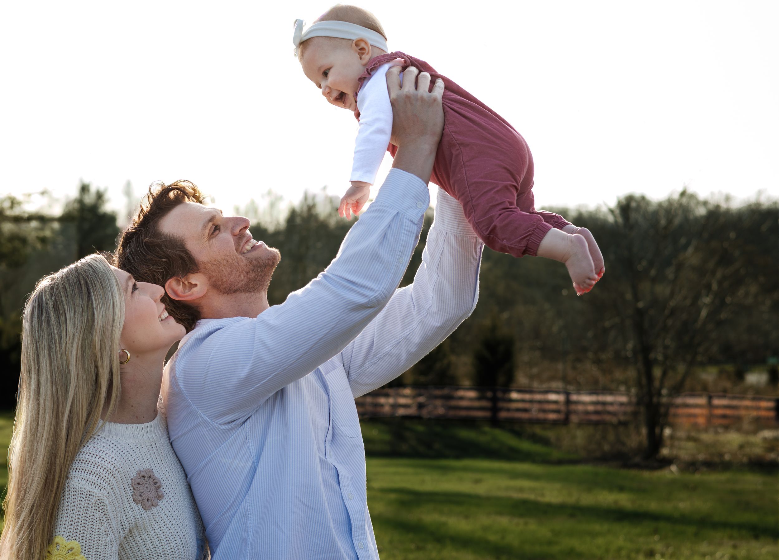 Happy parents lifting laughing baby in the air outdoors during a golden hour family photo session.