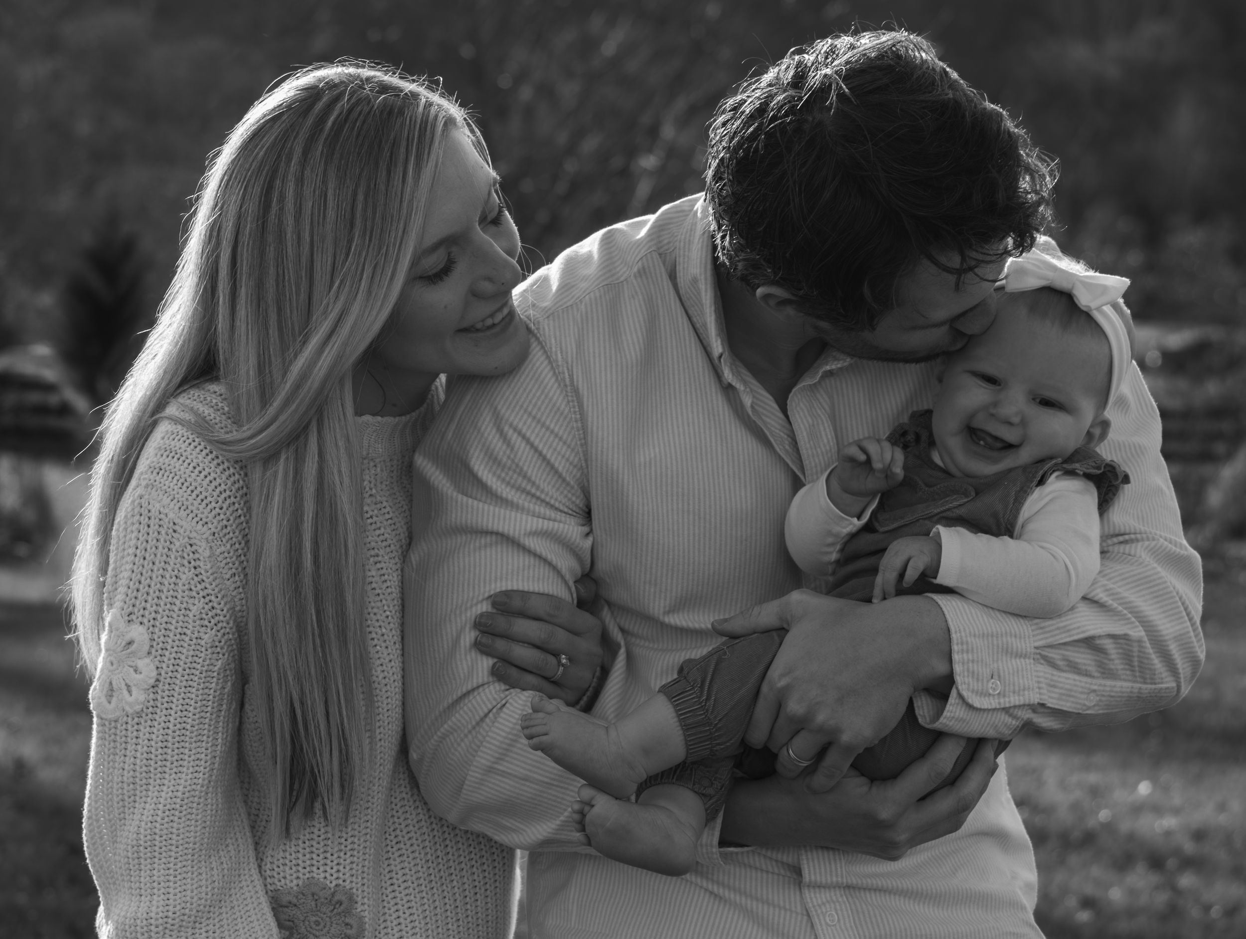 Black and white photo of loving parents kissing and cuddling their giggling baby outdoors together.