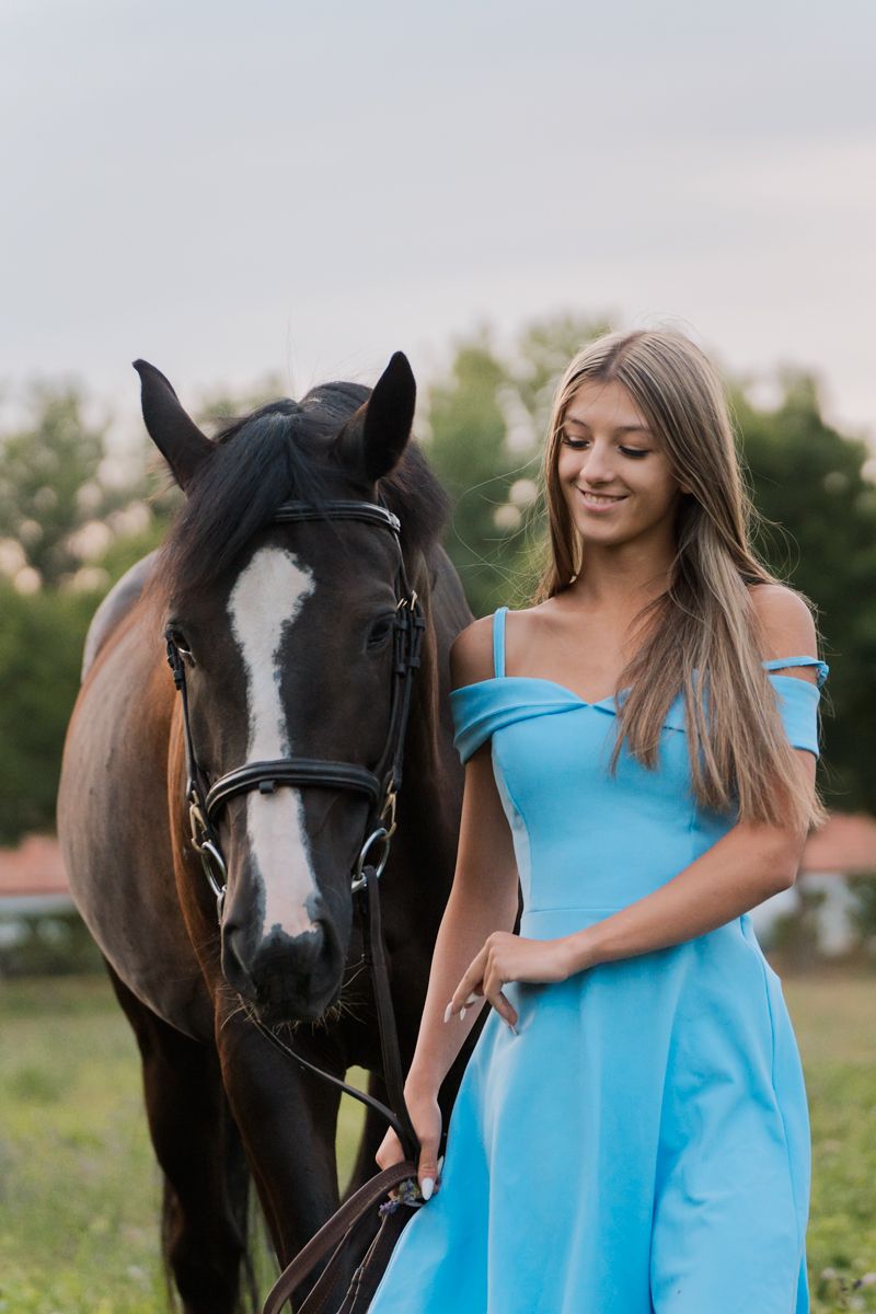 A beautiful horse with a white blaze stands next to a girl in a light blue off-shoulder dress in a grassy field during an equine photoshoot