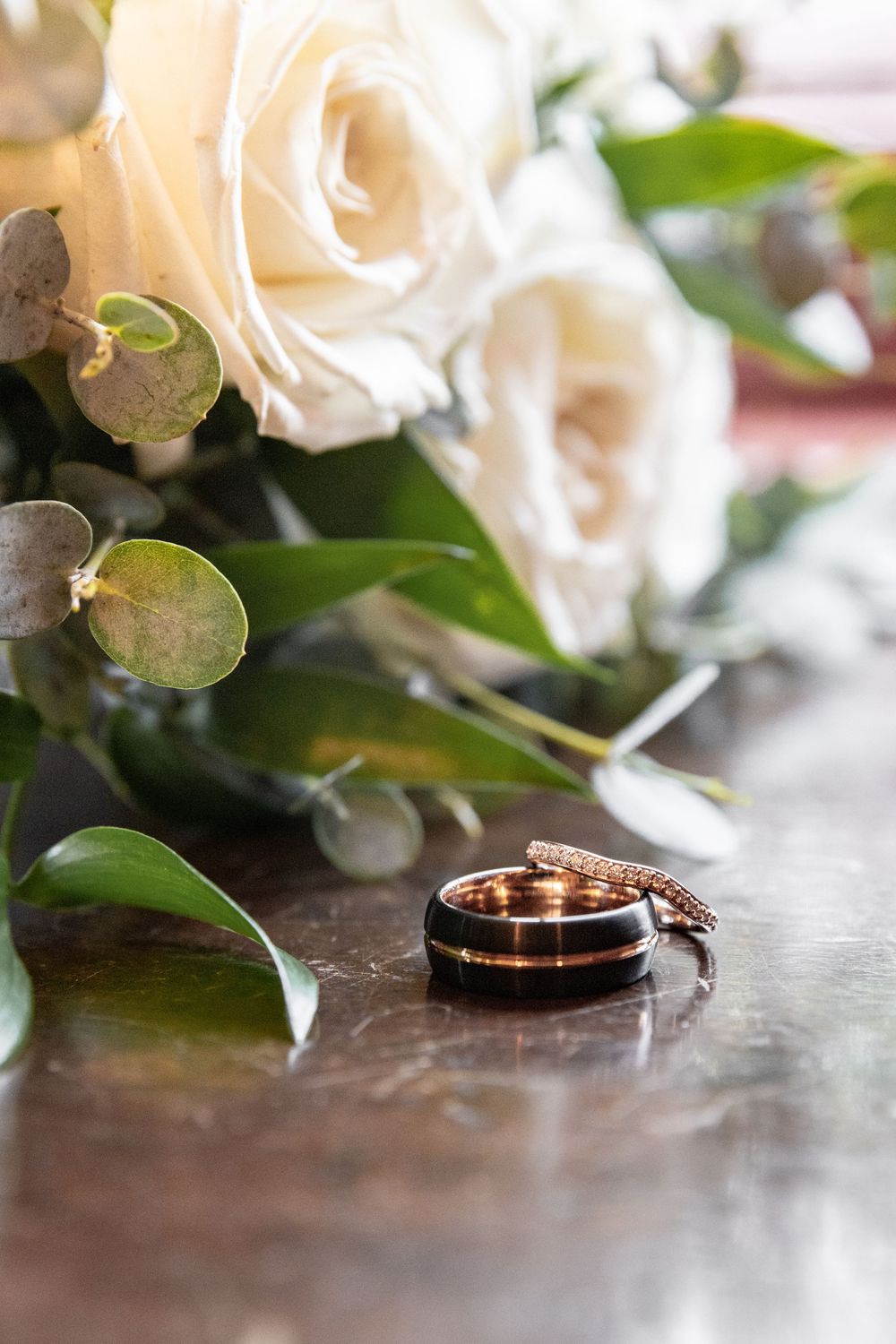 Gold wedding ring with black inlay rests beside white roses and eucalyptus leaves on dark wooden surface.