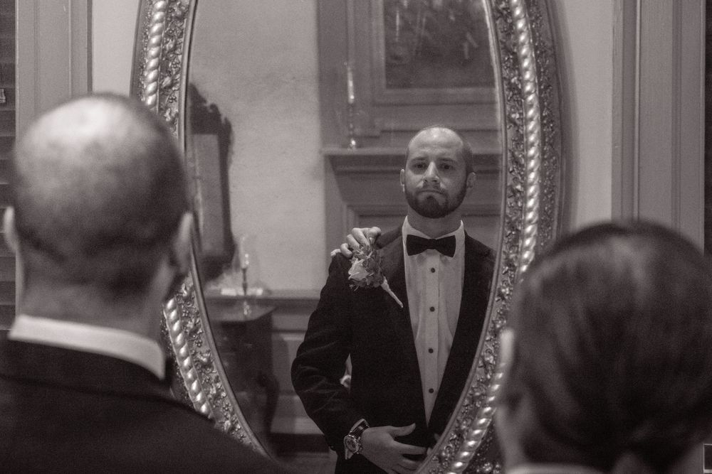 A black and white photograph shows several people in formal attire gathered around a large ornate mirror.