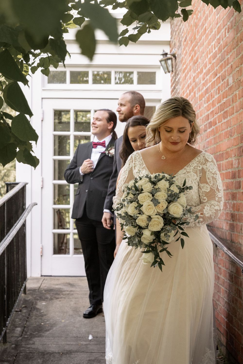 A bride in a lace wedding dress holds a white rose bouquet while walking down a brick-lined path.