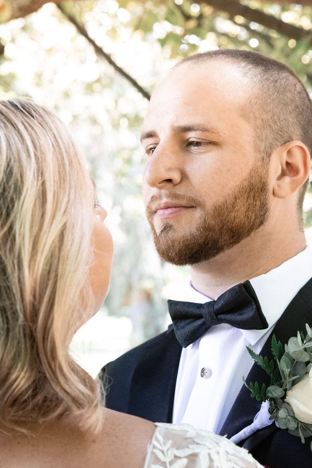 A couple in wedding attire share an intimate moment outdoors with soft natural lighting and greenery in the background.